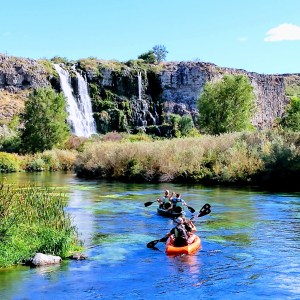 Kayakers paddle towards a waterfall in a scenic river surrounded by greenery and rocky cliffs.