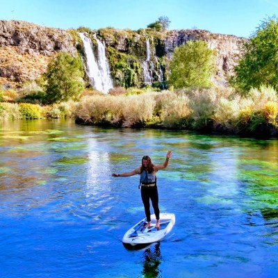 Person paddleboarding on a river with a waterfall and trees in the background.