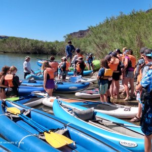 Group of people in life jackets with inflatable kayaks and paddleboards by a riverbank.