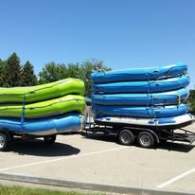 Two trailers loaded with stacked blue and green rafts in a parking lot under a clear blue sky.