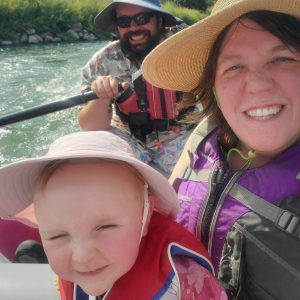 Three people in a raft on a sunny day, wearing hats and life vests, with a river and hills in the background.