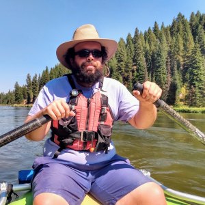 Person kayaking on a river wearing a hat and lifejacket, surrounded by pine trees.