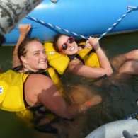 Two people in yellow life jackets enjoying water near a blue raft.