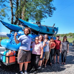 Group of seven people pose beside blue kayaks on a sunny day.