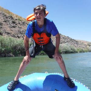 Person wearing life vest stands on inflatable raft in a river with hills in the background.