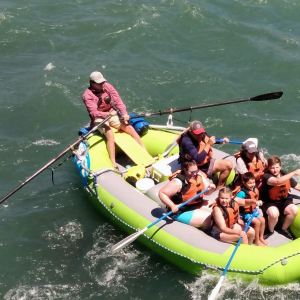 Group of people wearing life jackets rafting in a green inflatable boat on a river.