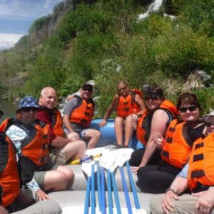 Group of people in life jackets on a raft by riverbank with greenery.