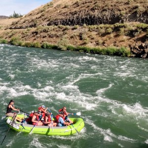 Group white water rafting in a green raft on a river, surrounded by hilly terrain.