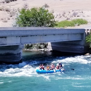 Group rafting on a river under a concrete bridge near desert landscape.