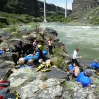 Group of people preparing for rafting by a rocky riverbank with inflatable rafts.
