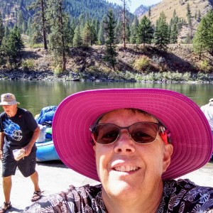 Person in pink hat taking a selfie by a river with trees and hills in the background.