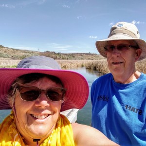 Two people wearing hats smiling near a river with grassy banks under a clear sky.