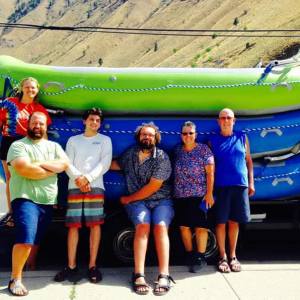 Six people standing in front of kayaks on a trailer, with a mountain in the background.