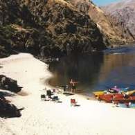 Sandy beach with chairs, kayaks, one person standing, and rocky hills in the background.