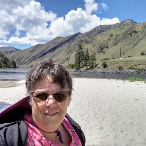 Person smiling with sunglasses at a riverside with mountains and blue sky in the background.
