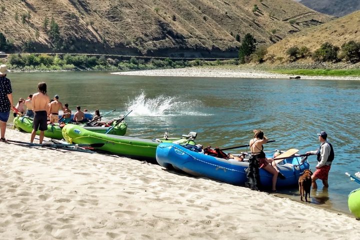 People with kayaks on a sandy riverbank, mountain and trees in the background.
