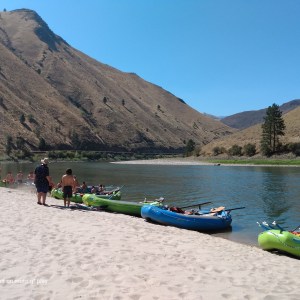 People with kayaks on a sandy riverbank beside grassy hills under a clear blue sky.