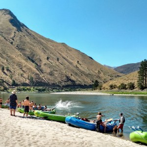 People and kayaks on a sandy riverbank with hills in the background.