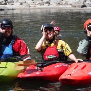 Three kayakers in helmets make playful gestures in a river.