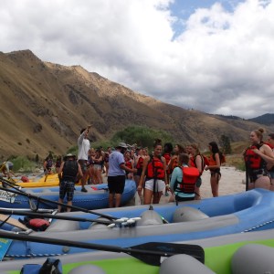 Group of people with life vests near rafts on a riverbank with hilly landscape in the background.