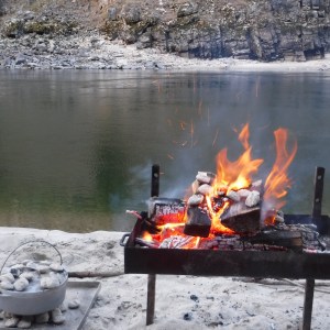 Campfire burning with rocks and wood by a river on a sandy shore.