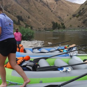 People preparing rafts on a riverbank, backed by hilly terrain.