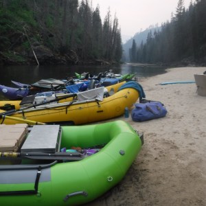 Inflatable rafts docked on a sandy riverbank with forested mountains in the background.