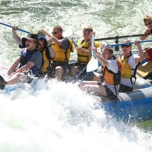Group of people white-water rafting in a blue inflatable raft, smiling and paddling in the rapids.